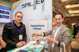 Двама участници на събитие, един мъж и една жена, пред щанд с надпис 'ендодонтско лечение'.