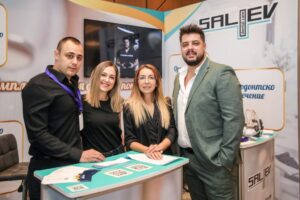 Четирима души, стоящи пред щанд на SALIEV на събитие, облечени в официални дрехи.