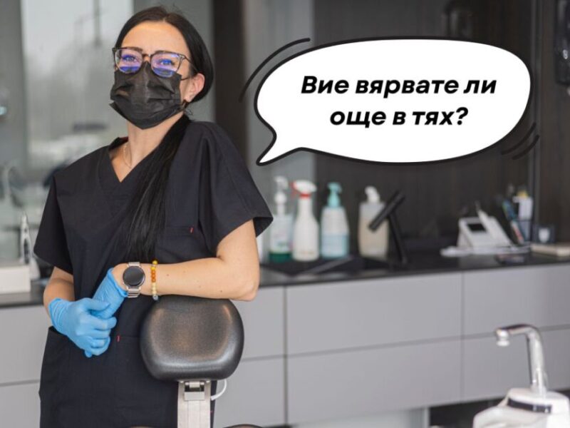 Зъболекар в черна униформа с маска и ръкавици, стояща в стоматологичен кабинет.
