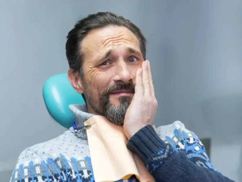 Мъж с израз на болка, държащ се за лицето в стоматологичен кабинет.