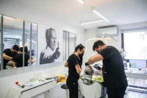 Двама стоматолози работят с пациент в стоматологичен кабинет с огледала и снимка на стената.