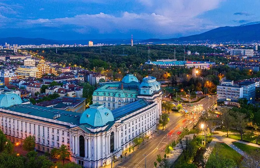 Панорамна гледка на София с осветени сгради и улици по време на залез.